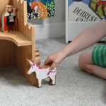 A close up of a Lanka Kade wooden unicorn figure in a child's hand. The Lanka Kade wooden toy castle can be seen in the background.