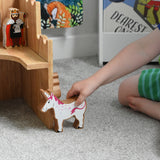 A close up of a Lanka Kade wooden unicorn figure in a child's hand. The Lanka Kade wooden toy castle can be seen in the background.