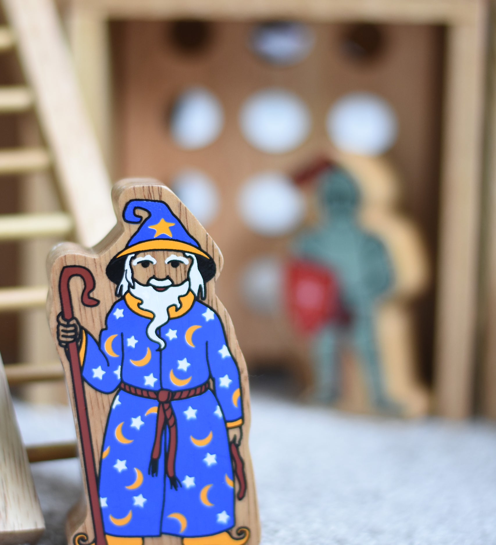 A Lanka Kade wooden toy wizard figure. The wizard has a blue coloured cloak with white and yellow stars and moon pattern withe a matching hat. The wizard is also holding a tall brown stick. 
