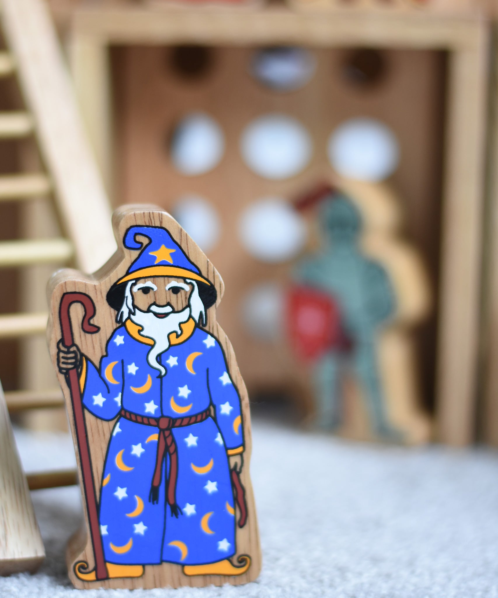A Lanka Kade wooden toy wizard figure. The wizard has a blue coloured cloak with white and yellow stars and moon pattern withe a matching hat. The wizard is also holding a tall brown stick. 