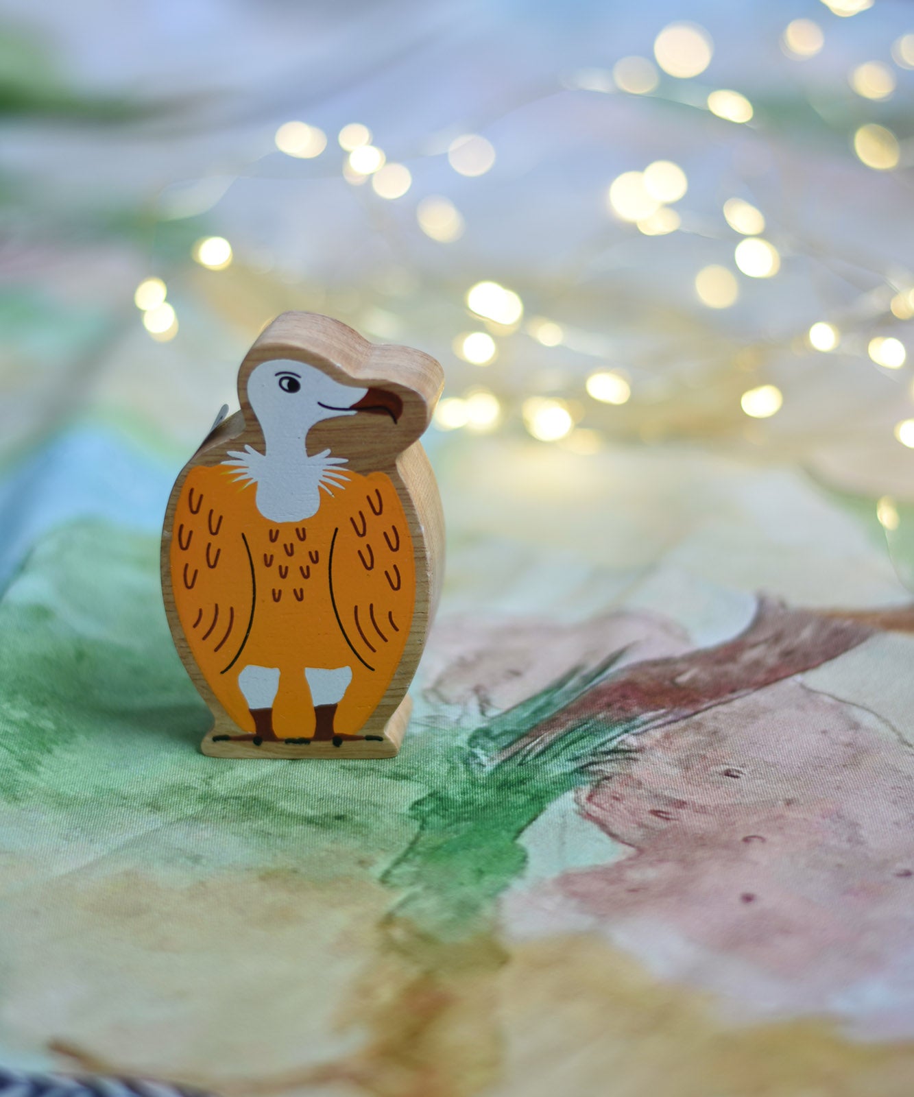 A Lanka Kade Wooden Yellow and White Vulture Toy placed on a Wonderie play cloth with fairy lights in the background. These wooden toy figures are part of a huge range of Lanka Kade fair trade, wooden toys available here at Babipur.