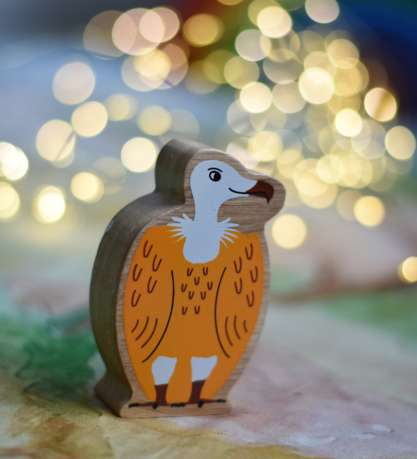 A Lanka Kade Wooden Yellow and White Vulture Toy placed on a Wonderie play cloth with fairy lights in the background. These wooden toy figures are part of a huge range of Lanka Kade fair trade, wooden toys available here at Babipur.