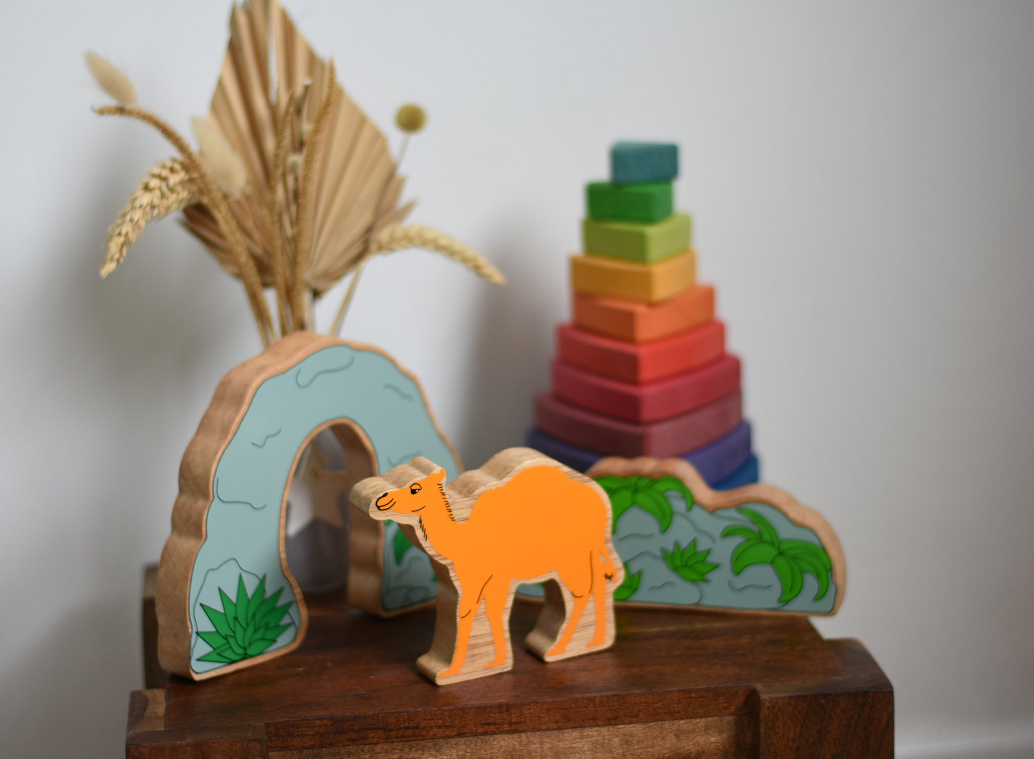 A Lanka Kade camel wooden toy figure placed on a small wooden table with the scenery pieces from the prehistoric play set. A Grimm's Wankel stacking toy can be seen in the background. These wooden toy figures are part of a huge range of Lanka Kade fair trade, wooden toys available here at Babipur.