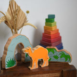 A Lanka Kade camel wooden toy figure placed on a small wooden table with the scenery pieces from the prehistoric play set. A Grimm's Wankel stacking toy can be seen in the background. These wooden toy figures are part of a huge range of Lanka Kade fair trade, wooden toys available here at Babipur.