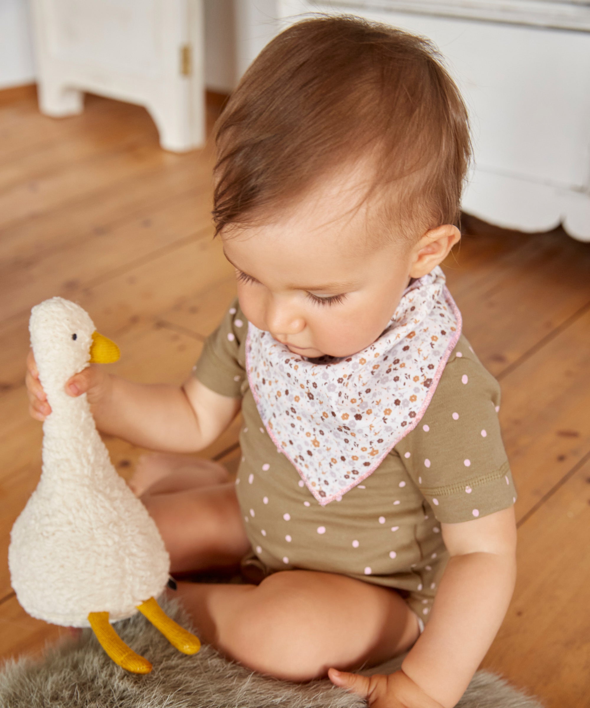 A little baby playing with a goose toy, and wearing the Lassig organic cotton bandana bib. 