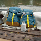 The Lassig kids big outdoor backpack in Adventure Blue, placed on a wooden surface outdoors next to the mini size. These children's backpacks are part of a wide range available at Babipur.