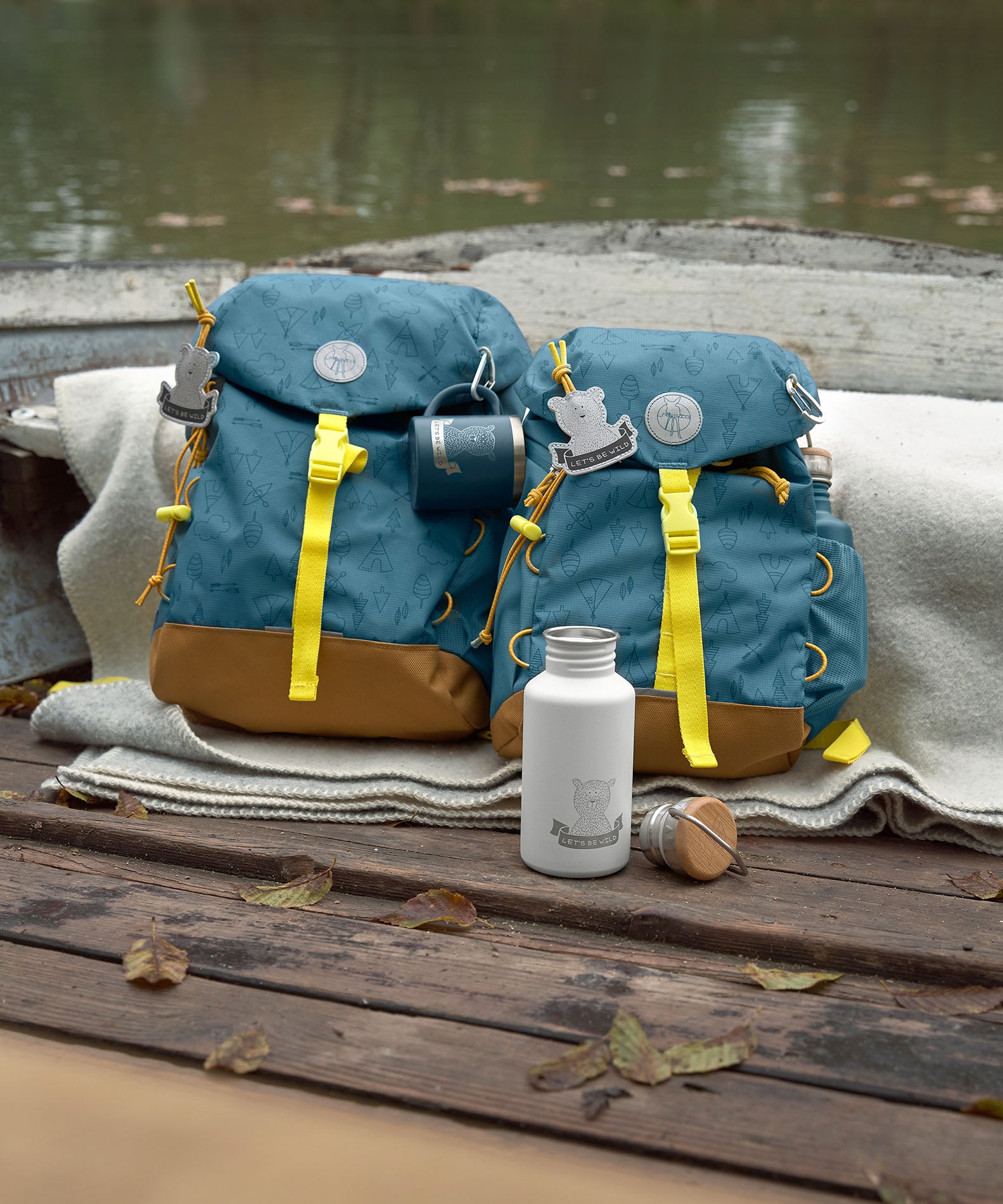 The Lassig kids big outdoor backpack in Adventure Blue, placed on a wooden surface outdoors next to the mini size. These children's backpacks are part of a wide range available at Babipur.