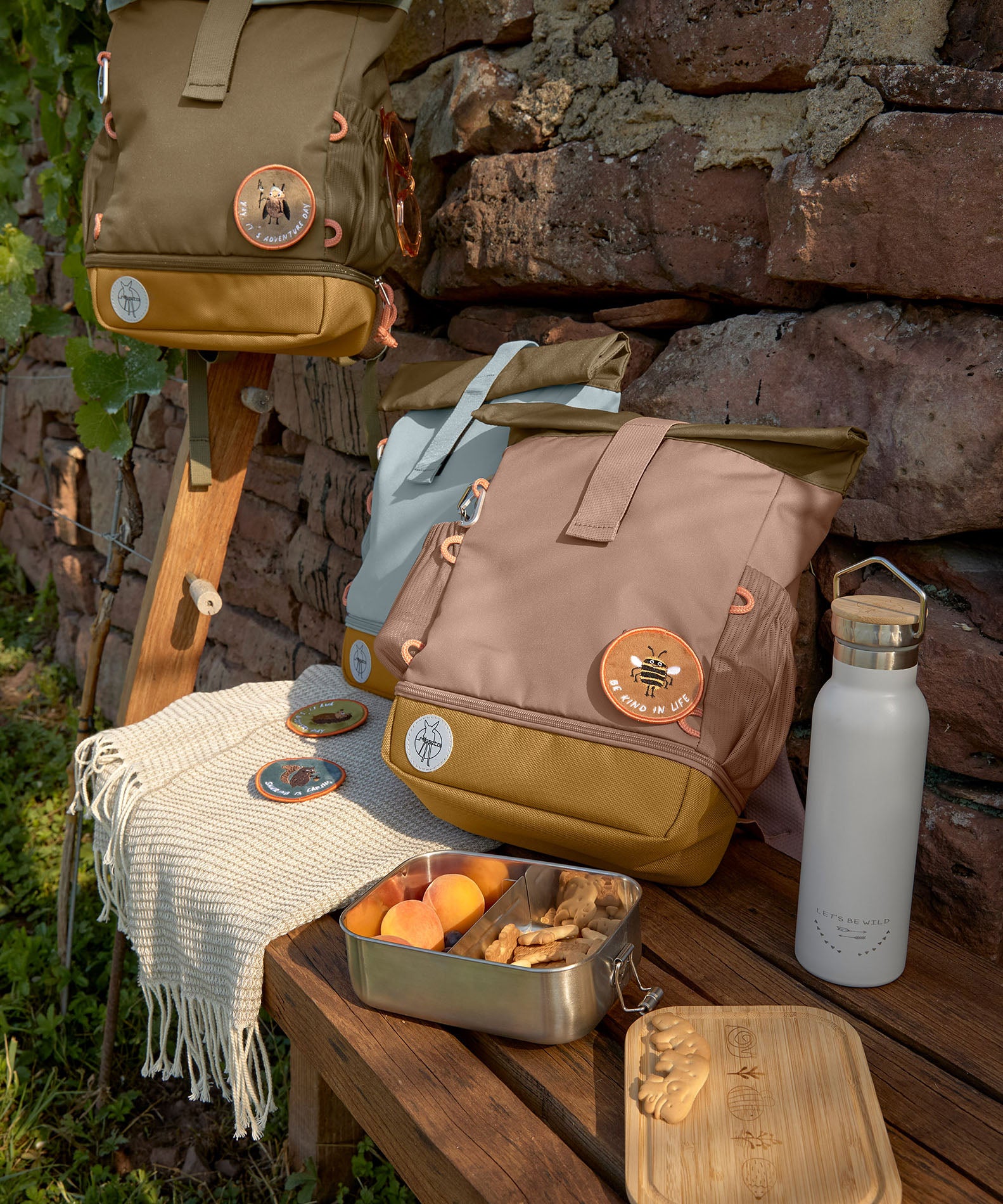The Lassig Mini Rolltop Backpack in a Nature Hazelnut colour placed on a bench outdoors. Other colours of the bag can be seen in the background. This children's bag is part of a wide range of backpacks and accessories available at Babipur. 