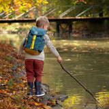 A child wearing the Lassig Kids Big Outdoor Backpack with an Adventure Blue design. The print features tents, trees, and clouds, and a neon green front strap. These children's backpacks are part of a wide range available at Babipur.