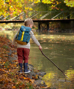 A child wearing the Lassig Kids Big Outdoor Backpack with an Adventure Blue design. The print features tents, trees, and clouds, and a neon green front strap. These children's backpacks are part of a wide range available at Babipur.