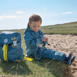 A child sitting on the ground outdoors with the Lassig Kids Big Outdoor Backpack with an Adventure Blue design placed on the ground next to them. The print features tents, trees, and clouds, and a neon green front strap. These children's backpacks are part of a wide range available at Babipur.