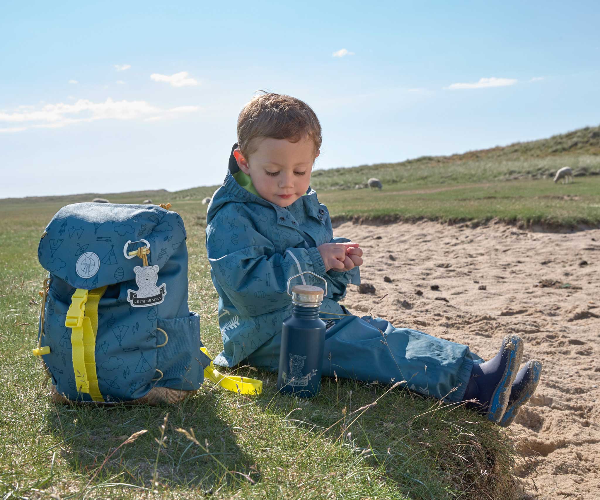 A child sitting on the ground outdoors with the Lassig Kids Big Outdoor Backpack with an Adventure Blue design placed on the ground next to them. The print features tents, trees, and clouds, and a neon green front strap. These children's backpacks are part of a wide range available at Babipur.