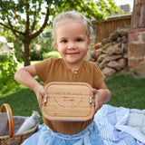 A child holding a Lassig Stainless Steel Bamboo Lunchbox with a Garden Explorer design. This stainless steel lunchbox is part of a wide range of reusable food containers available at Babipur. 