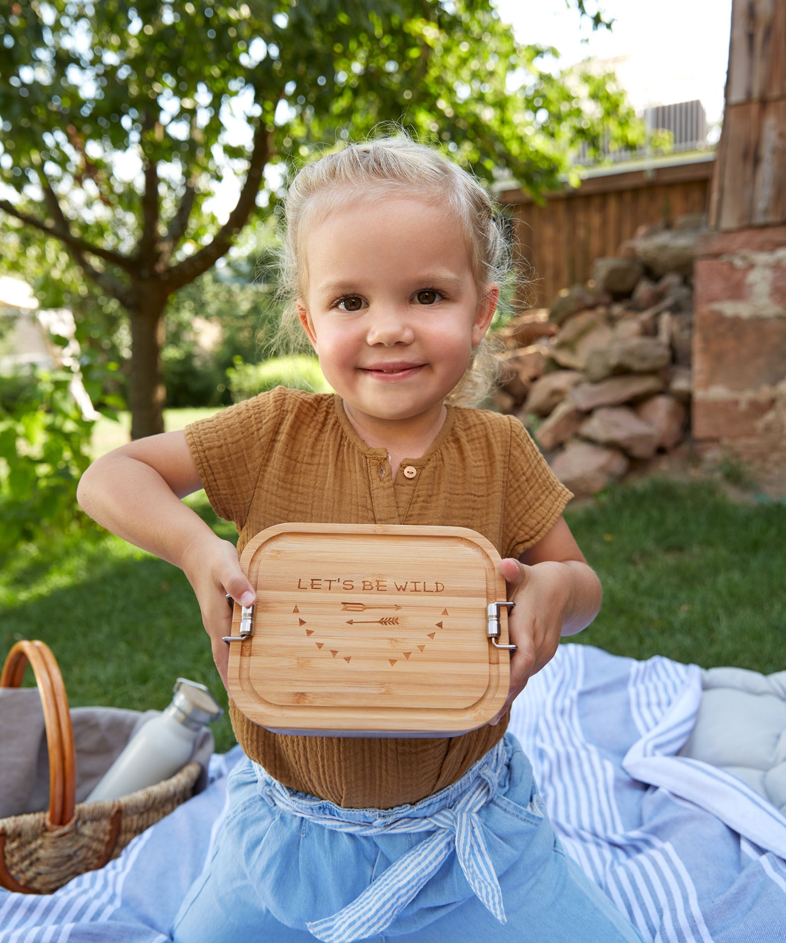 A child holding a Lassig Stainless Steel Bamboo Lunchbox with a Garden Explorer design. This stainless steel lunchbox is part of a wide range of reusable food containers available at Babipur. 