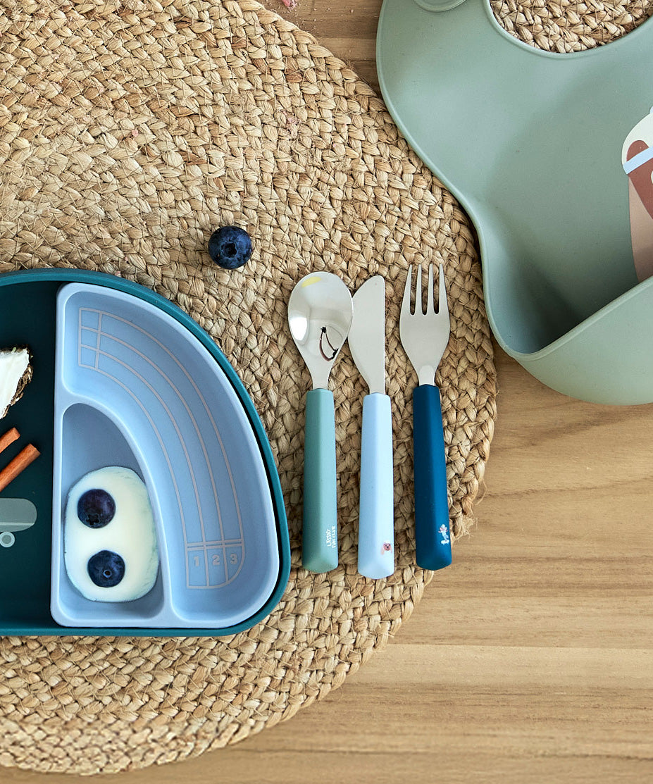 The Lassig Cutlery with Silicone Handle with a Tiny Team Dog design shown on a table ready to be used. These green and blue coloured children's cutlery are part of a wide range of tabelware avaialable at Babipur.