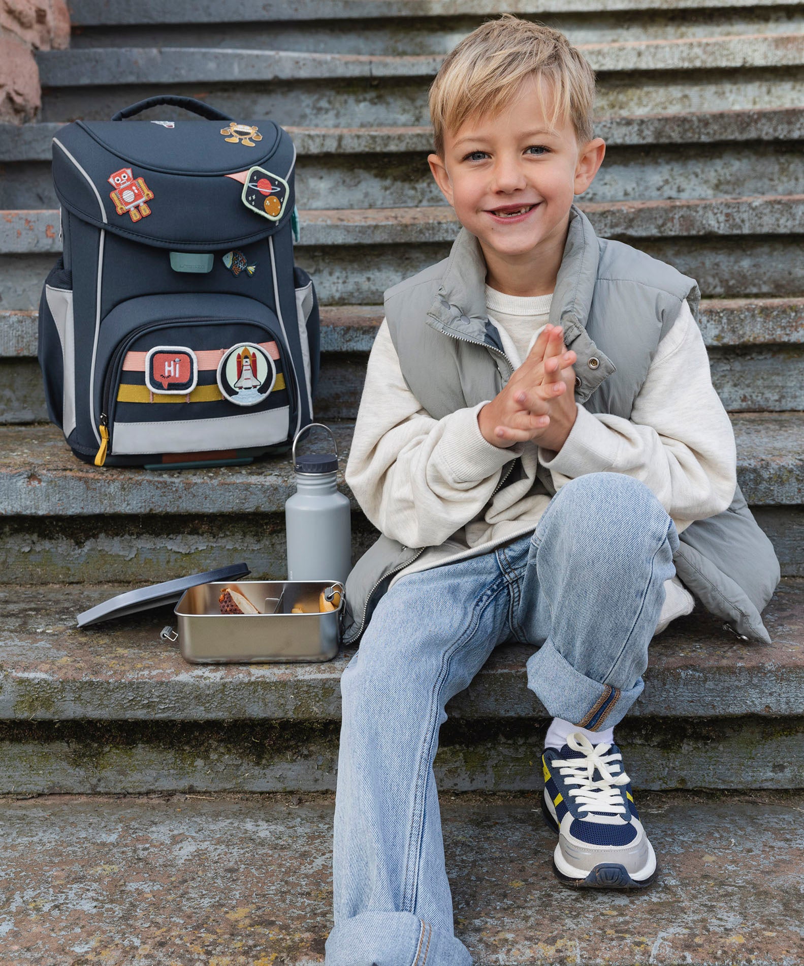 A child sitting on steps next to a filled Lassig Stainless Steel Lunchbox with a Unique Dark Blue lid. This lunch box has a removable divider. 