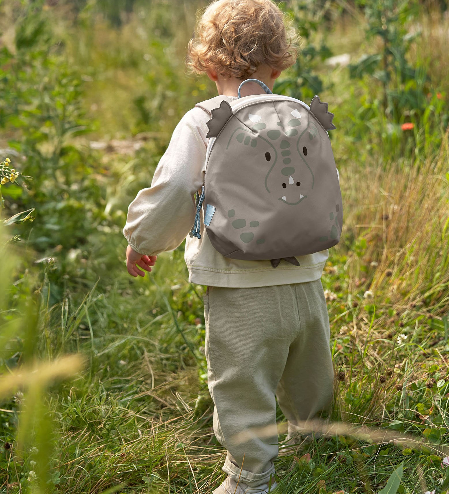 A child walking through a grassy gfield and wearing the Lassig kids tiny dragon backpack