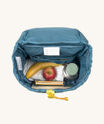 A view of the interior of the Lassig Kids Big Outdoor Backpack with an Adventure Blue design. A bottle, fruit and some books can be seen inside. The print features tents, trees, and clouds, and a neon green front strap. These children's backpacks are part of a wide range available at Babipur.