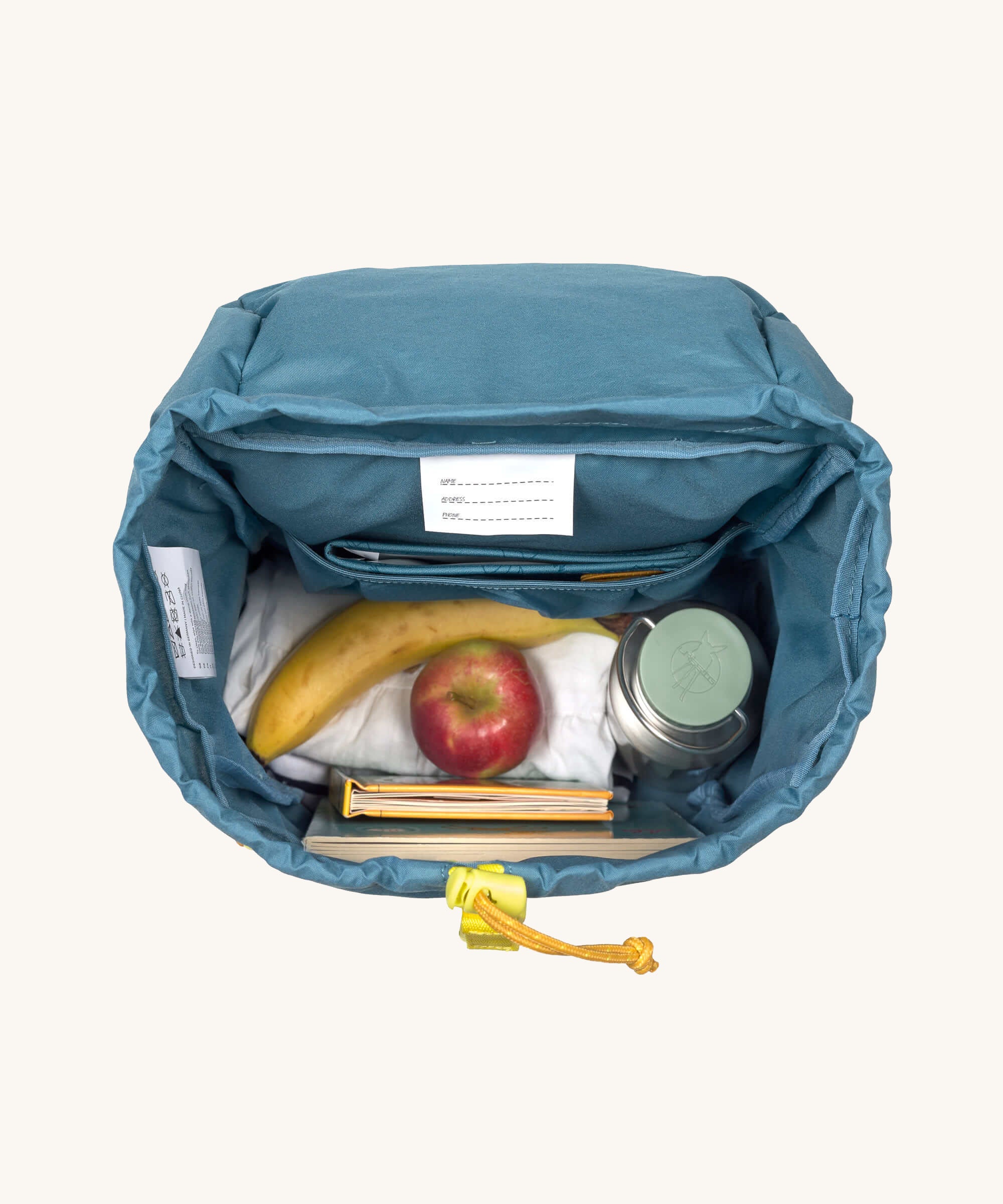 A view of the interior of the Lassig Kids Big Outdoor Backpack with an Adventure Blue design. A bottle, fruit and some books can be seen inside. The print features tents, trees, and clouds, and a neon green front strap. These children's backpacks are part of a wide range available at Babipur.