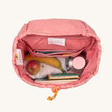 A top down viw of the interior of the Lassig Kids Big Outdoor Backpack with an Adventure rose pink design with items of clothing, fruit, books and a bottle inside. The print features tents, trees, and clouds, and a pink front strap. These children's backpacks are part of a wide range available at Babipur.