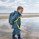 A child walking along a beach wearing the Lassig Kids Big Outdoor Backpack with an Adventure Blue design. The print features tents, trees, and clouds, and a neon green front strap. These children's backpacks are part of a wide range available at Babipur.