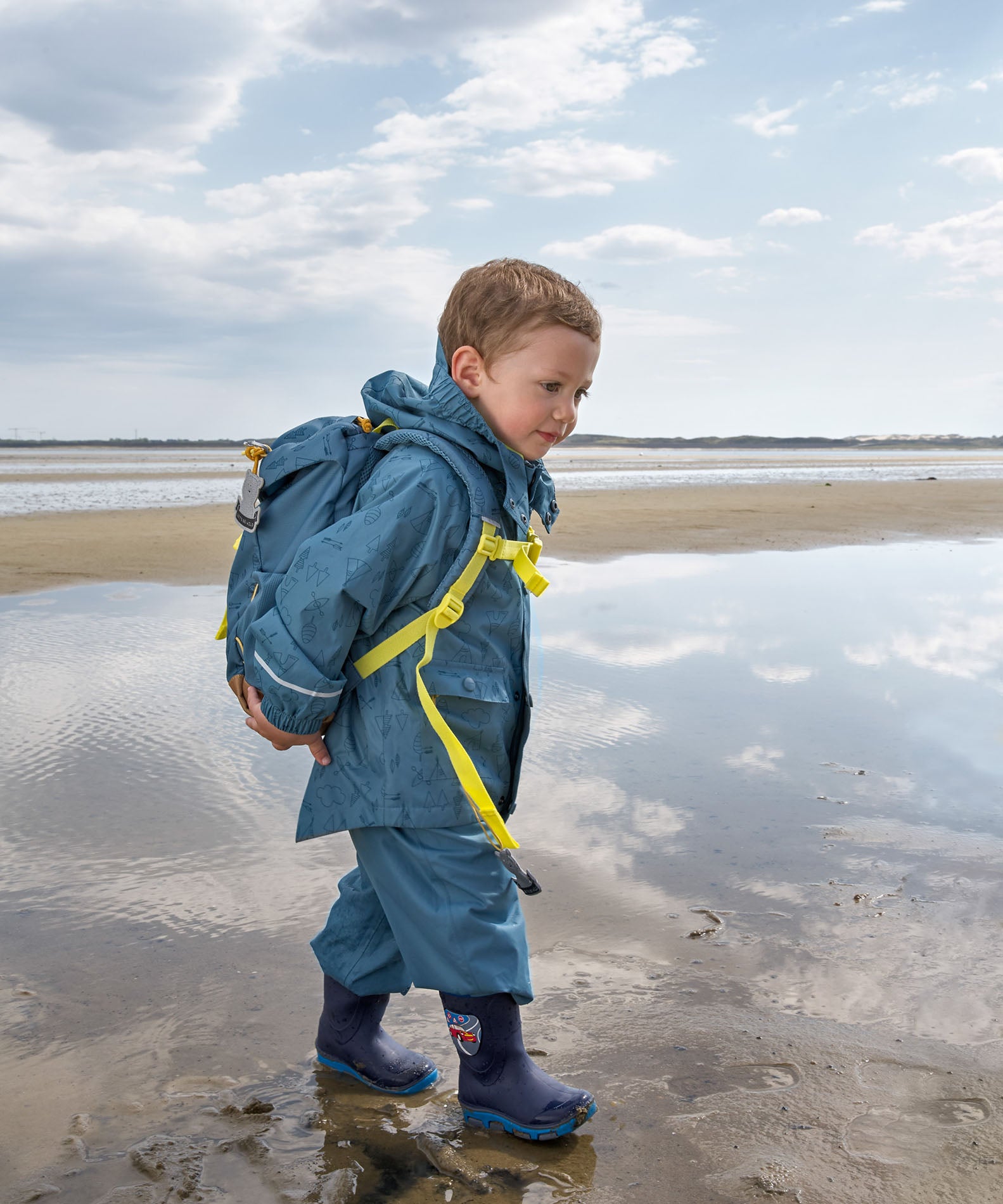 A child walking along a beach wearing the Lassig Kids Big Outdoor Backpack with an Adventure Blue design. The print features tents, trees, and clouds, and a neon green front strap. These children's backpacks are part of a wide range available at Babipur.