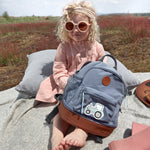 A little girl in sunglasses holding her Lassig kids mini backpack with a tractor on the front. 