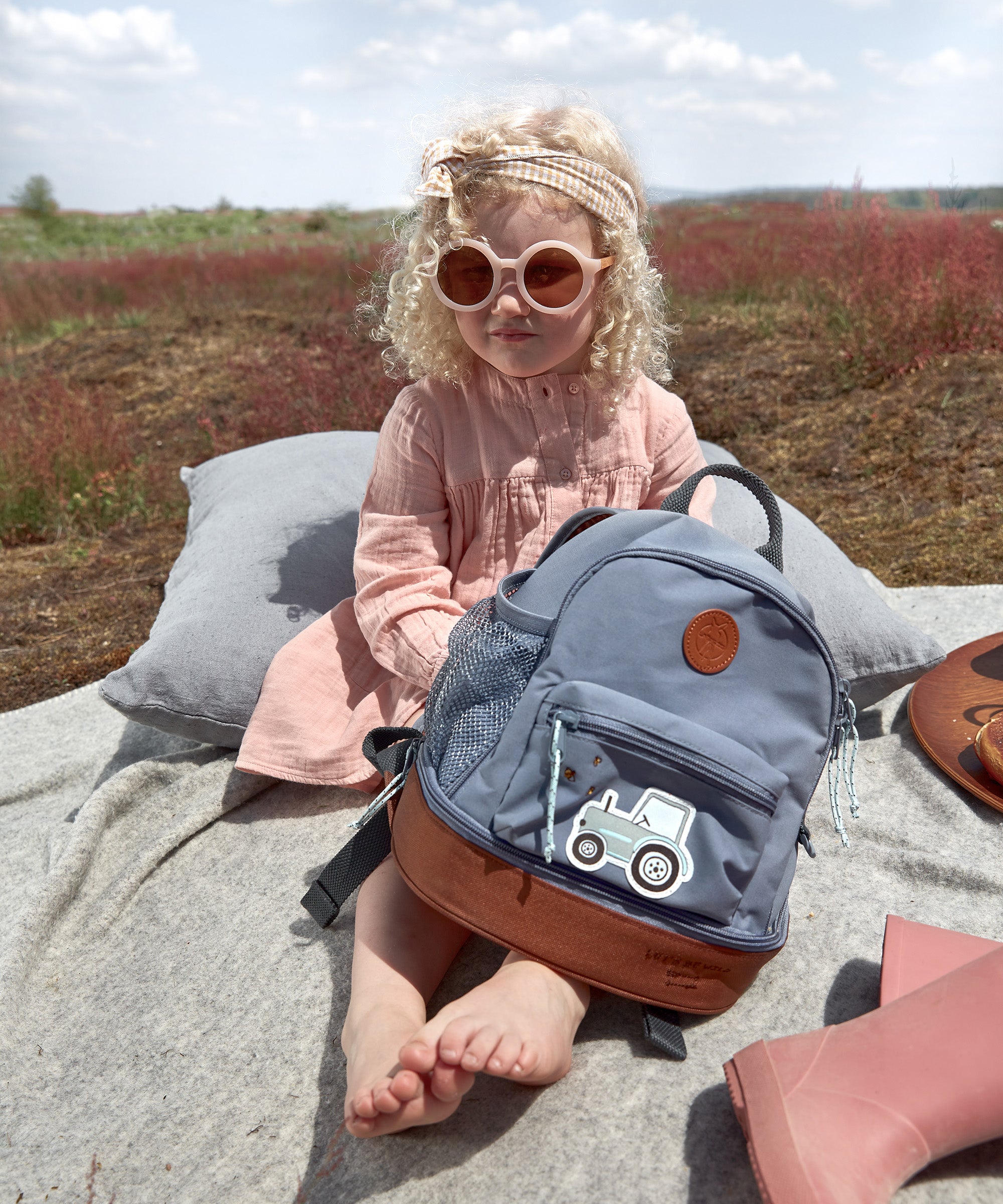 A little girl in sunglasses holding her Lassig kids mini backpack with a tractor on the front. 