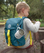 A little boy wearing the Lassig kids mini outdoor backpack in Adventure Blue, with a camping mug attached to it.