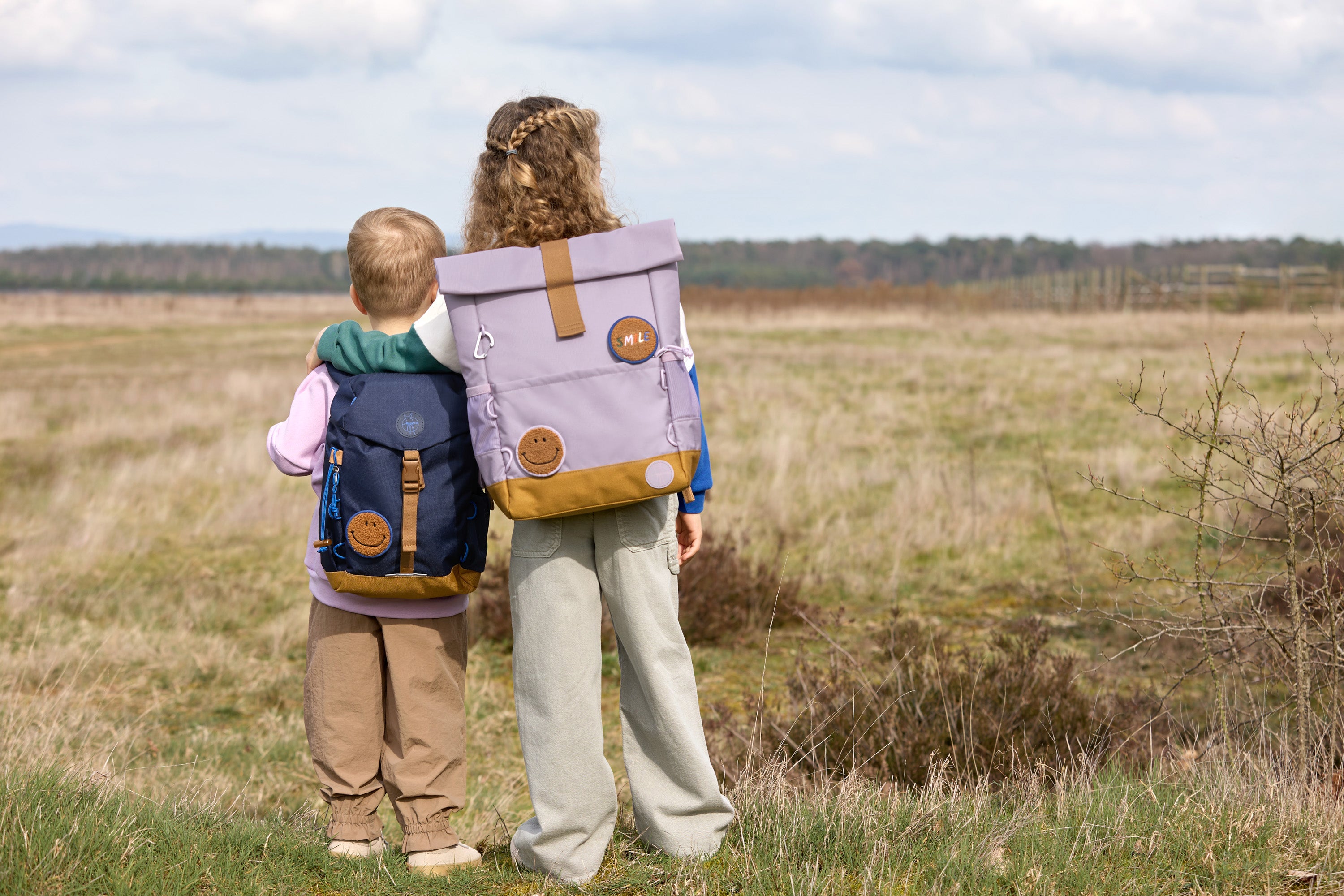 Two children wearing Lassig kids backpacks in a field. 