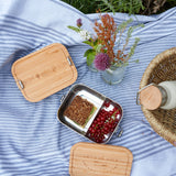 The Lassig Stainless Steel Bamboo Lunchbox with a Garden Explorer design shown open and filled with food. This stainless steel lunchbox is part of a wide range of reusable food containers available at Babipur. 