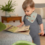 A toddler wearing the Lassig organic cotton baby bib in a Little Fox print, and reading a book. 