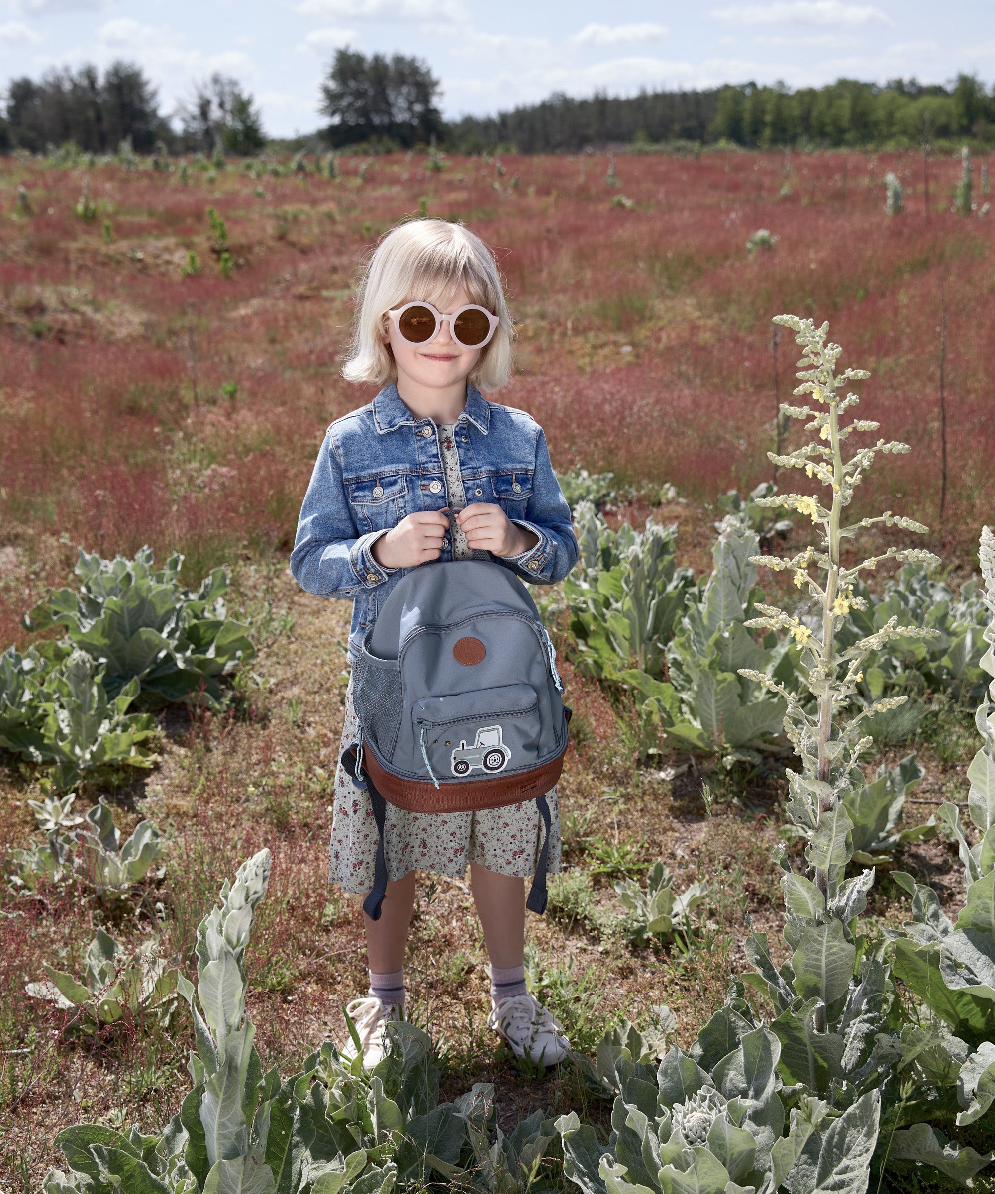 A girl in a sunny field, holding her Lassig mini backpack in blue with a tractor on it.