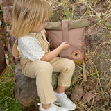 A child opening the roll top on the Lassig Mini Rolltop Backpack in a Nature Hazelnut colour. This children's bag is part of a wide range of backpacks and accessories available at Babipur. 