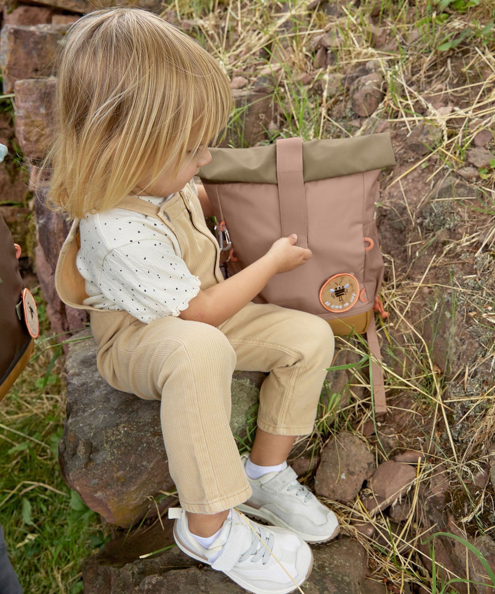 A child opening the roll top on the Lassig Mini Rolltop Backpack in a Nature Hazelnut colour. This children's bag is part of a wide range of backpacks and accessories available at Babipur. 