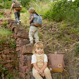 A child sitting on stone steps outdoors with a Lassig Mini Rolltop Backpack in a Nature Hazelnut colour on the step next to them. This children's bag is part of a wide range of backpacks and accessories available at Babipur. 
