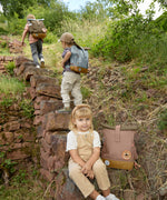 A child sitting on stone steps outdoors with a Lassig Mini Rolltop Backpack in a Nature Hazelnut colour on the step next to them. This children's bag is part of a wide range of backpacks and accessories available at Babipur. 
