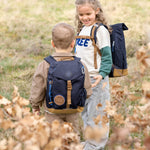 Two children wearing Lassig mini backpacks in navy blue, in a green field. 