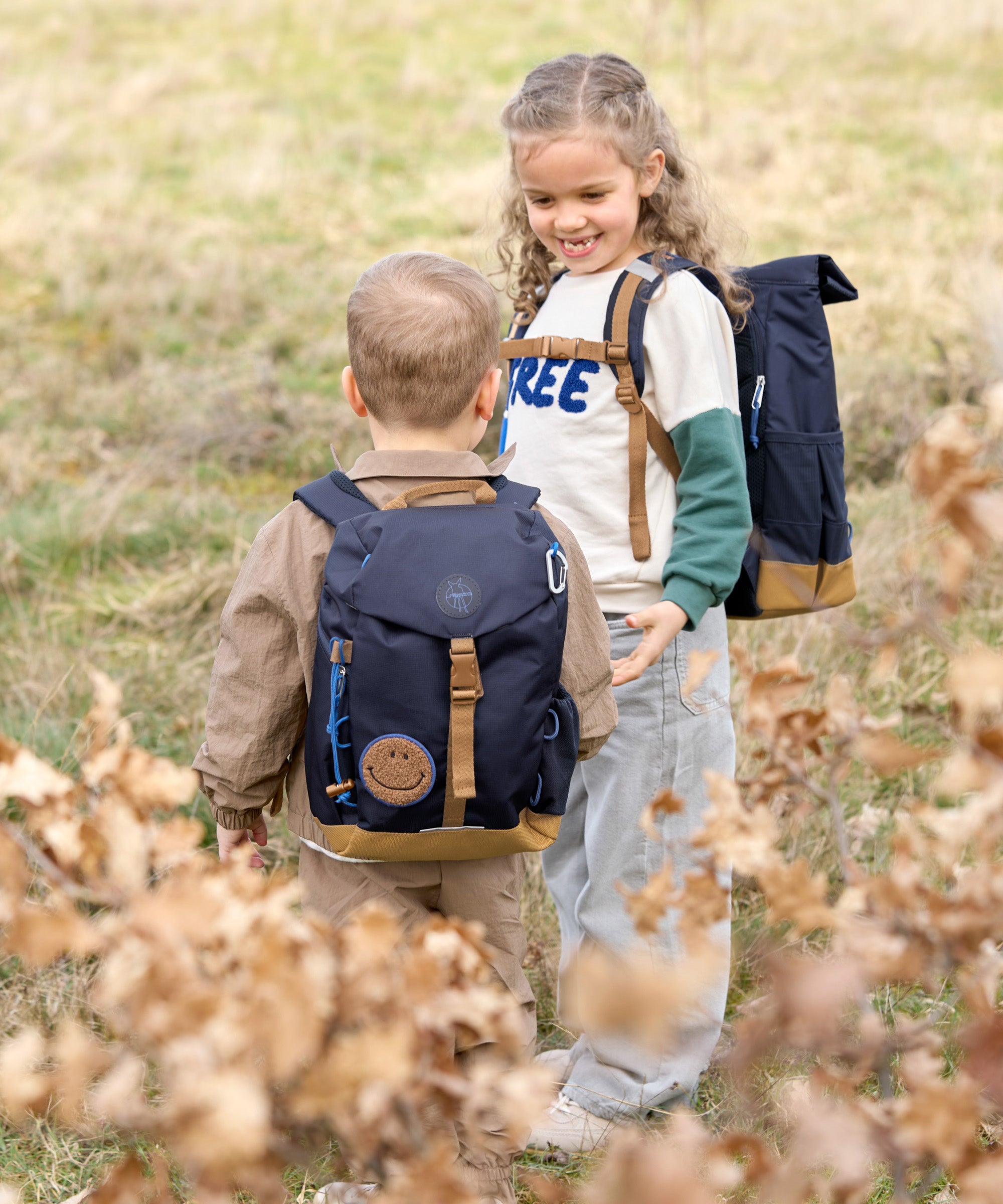 Two children wearing Lassig mini backpacks in navy blue, in a green field. 
