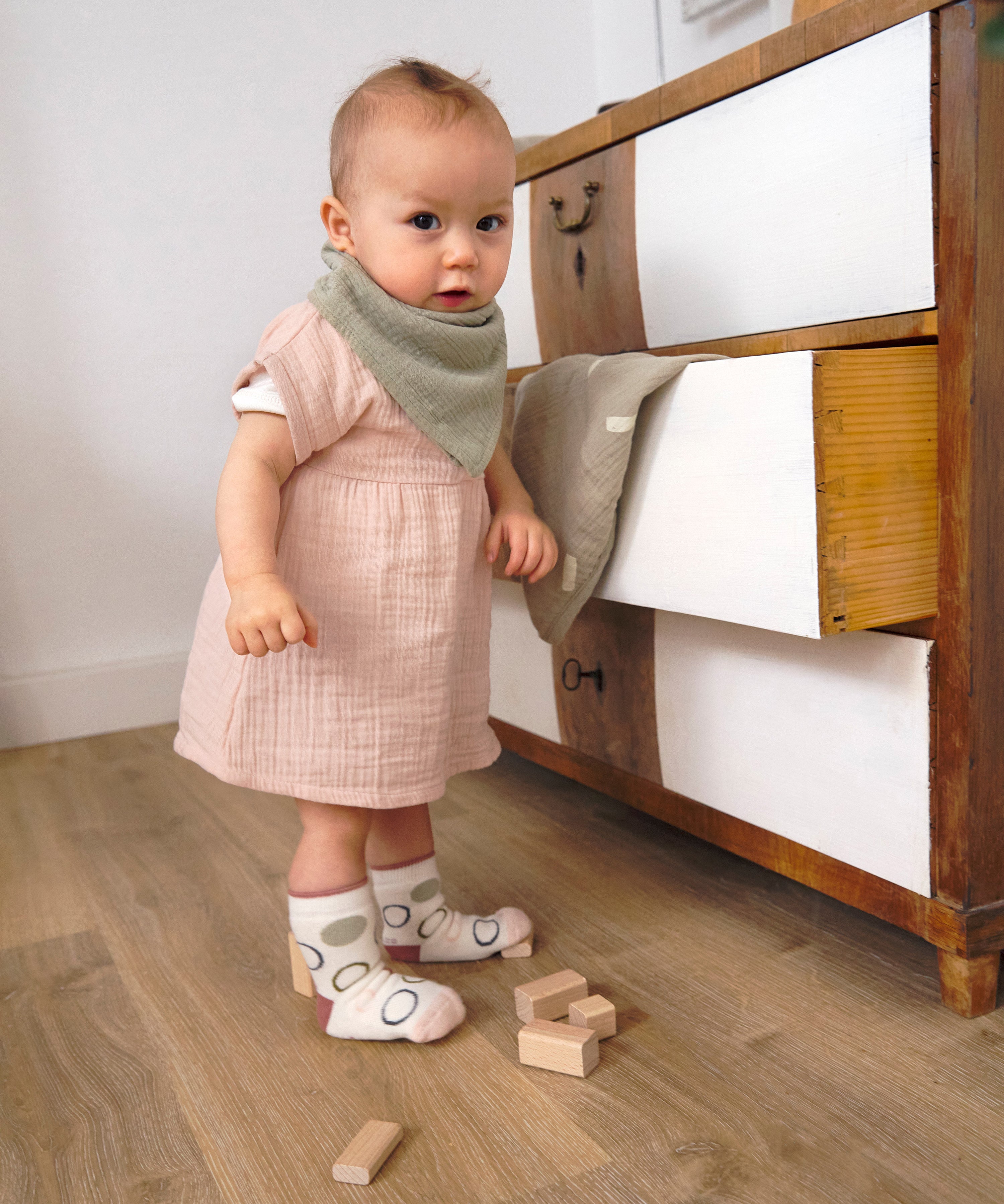 A baby wearing the Lassig organic cotton muslin dress with a bandana dribble bib.