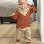 A little toddler wearing the Lassig organic cotton muslin shirt for babies and toddlers, in a bright orange rust colour.
