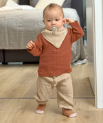 A little toddler wearing the Lassig organic cotton muslin shirt for babies and toddlers, in a bright orange rust colour.