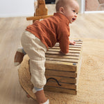 A baby climbing and wearing the Lassig organic cotton muslin shirt for babies and toddlers, in a bright orange rust colour.