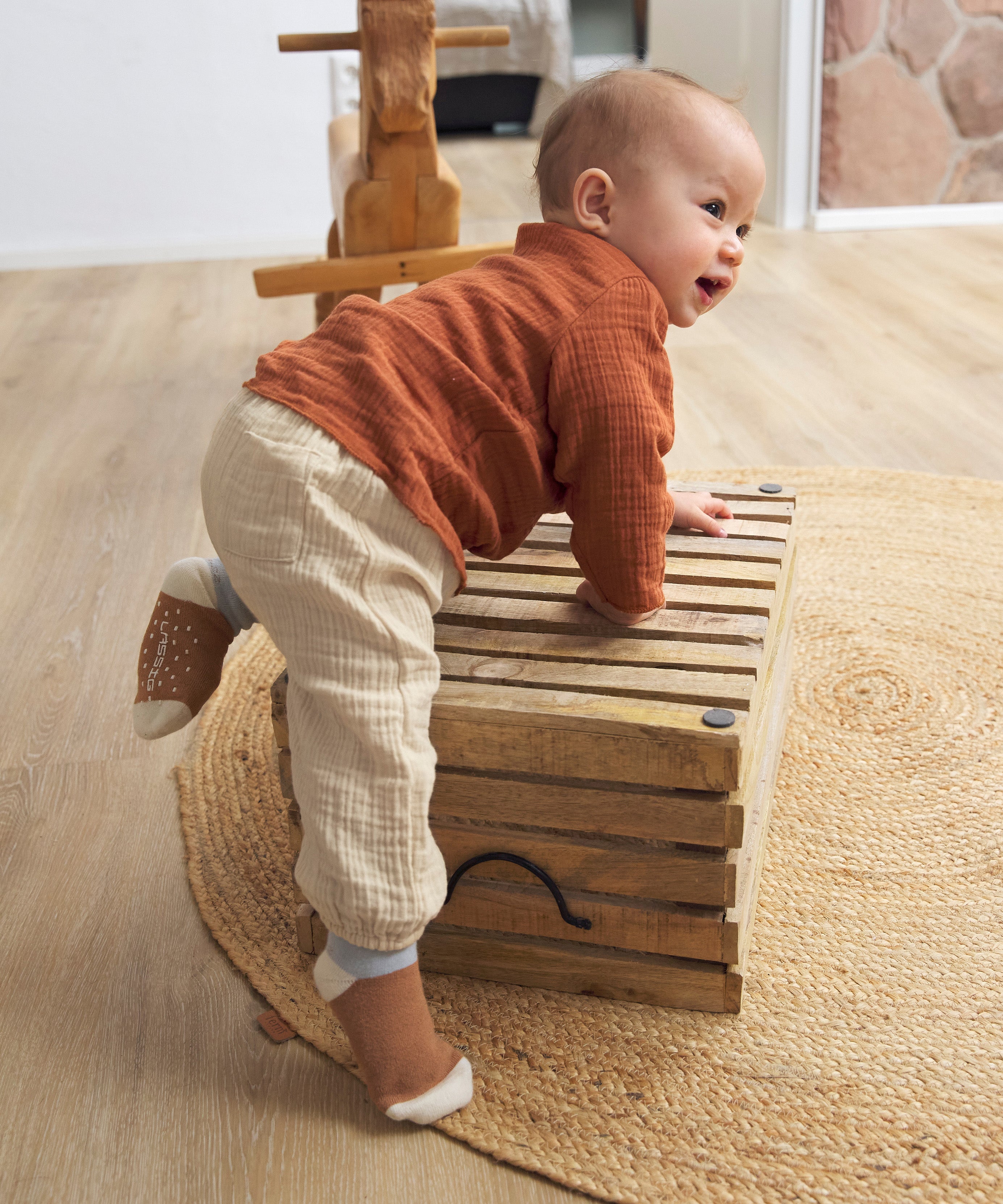 A baby climbing and wearing the Lassig organic cotton muslin shirt for babies and toddlers, in a bright orange rust colour.