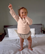 A baby standing on a bed, wearing the Lassig organic cotton muslin long sleeved shirt for babies and toddlers in a beautiful pale powder pink colour