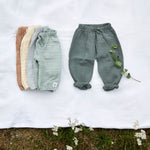 Different coloured Lassig organic cotton muslin baby pants, laid out on a blanket in a field. 
