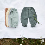 Different coloured Lassig organic cotton muslin baby pants, laid out on a blanket in a field. 