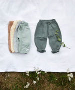 Different coloured Lassig organic cotton muslin baby pants, laid out on a blanket in a field. 
