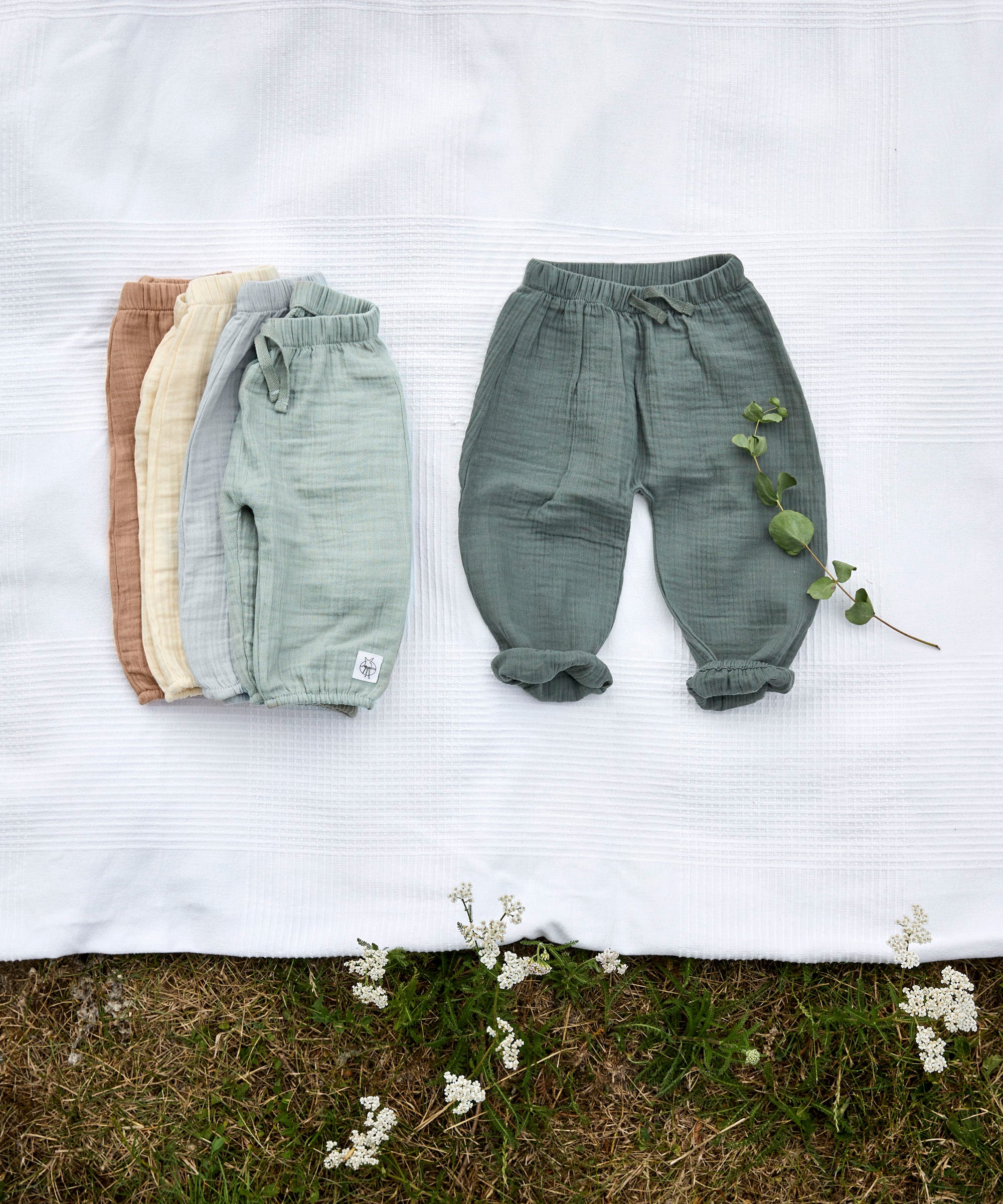 Different coloured Lassig organic cotton muslin baby pants, laid out on a blanket in a field. 