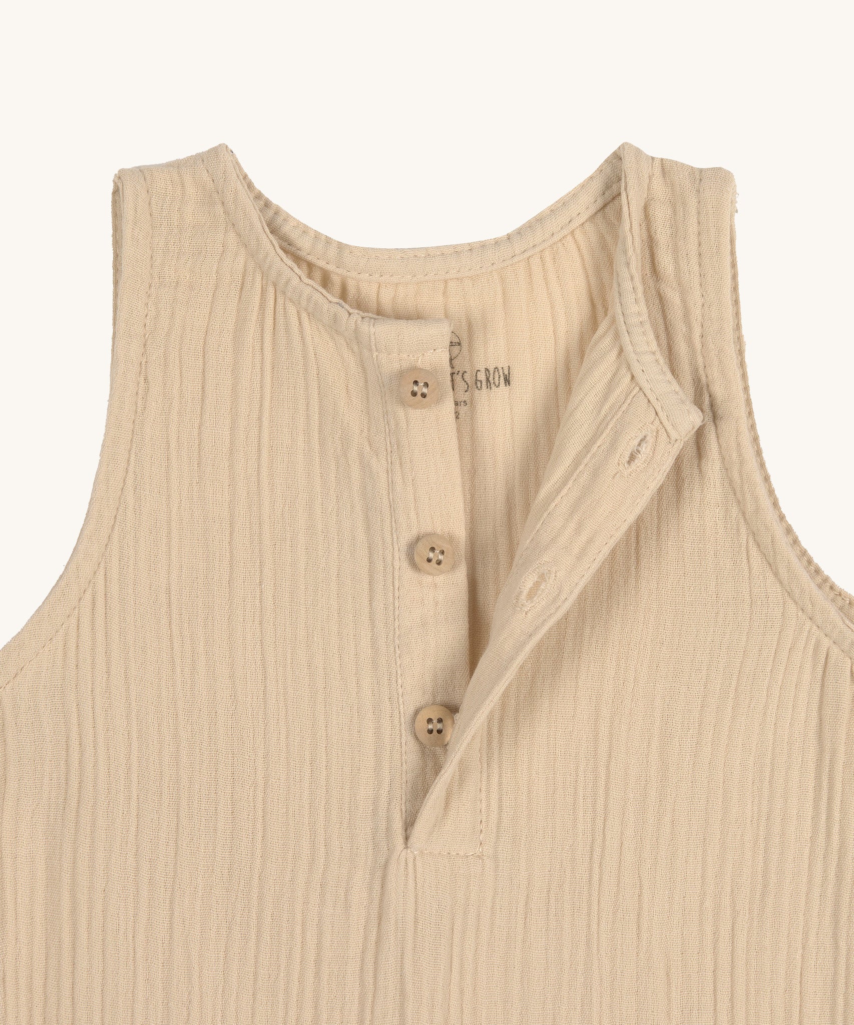 A closeup of the Lassig organic cotton muslin baby and toddler jumpsuit in a natural cream colour, showing the three cream buttons at the neck.